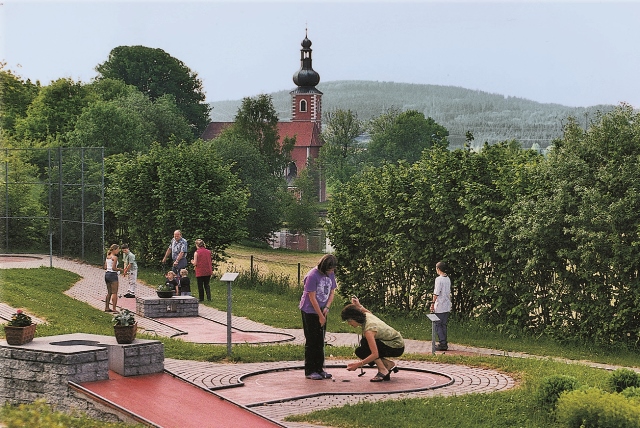 Panoramabild des Minigolfplatz mit der Kirche im Hintergrund und einigen Menschen, die Minigolf spielen.