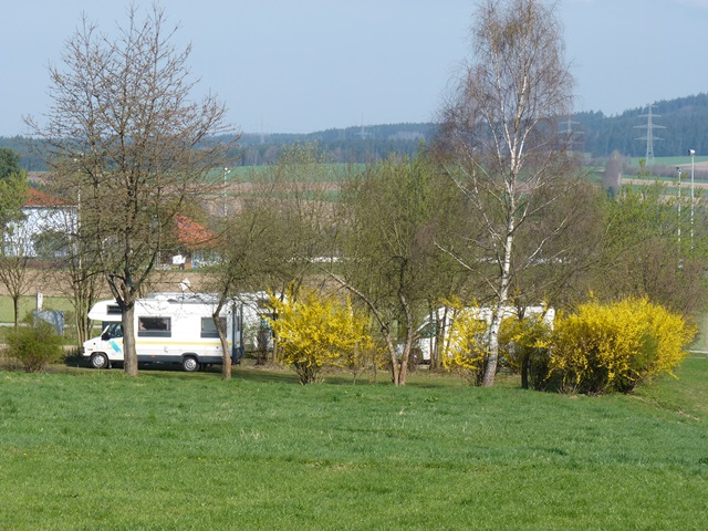 Mehrere Wohnmobile auf einem Parkplatz hinter Bäumen