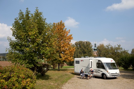 Ein Wohnmobil auf einem Naturparkplatz mit zwei Campern an einem Tisch. Im Hintergrund sieht man die Wieskirche Moosbachs.