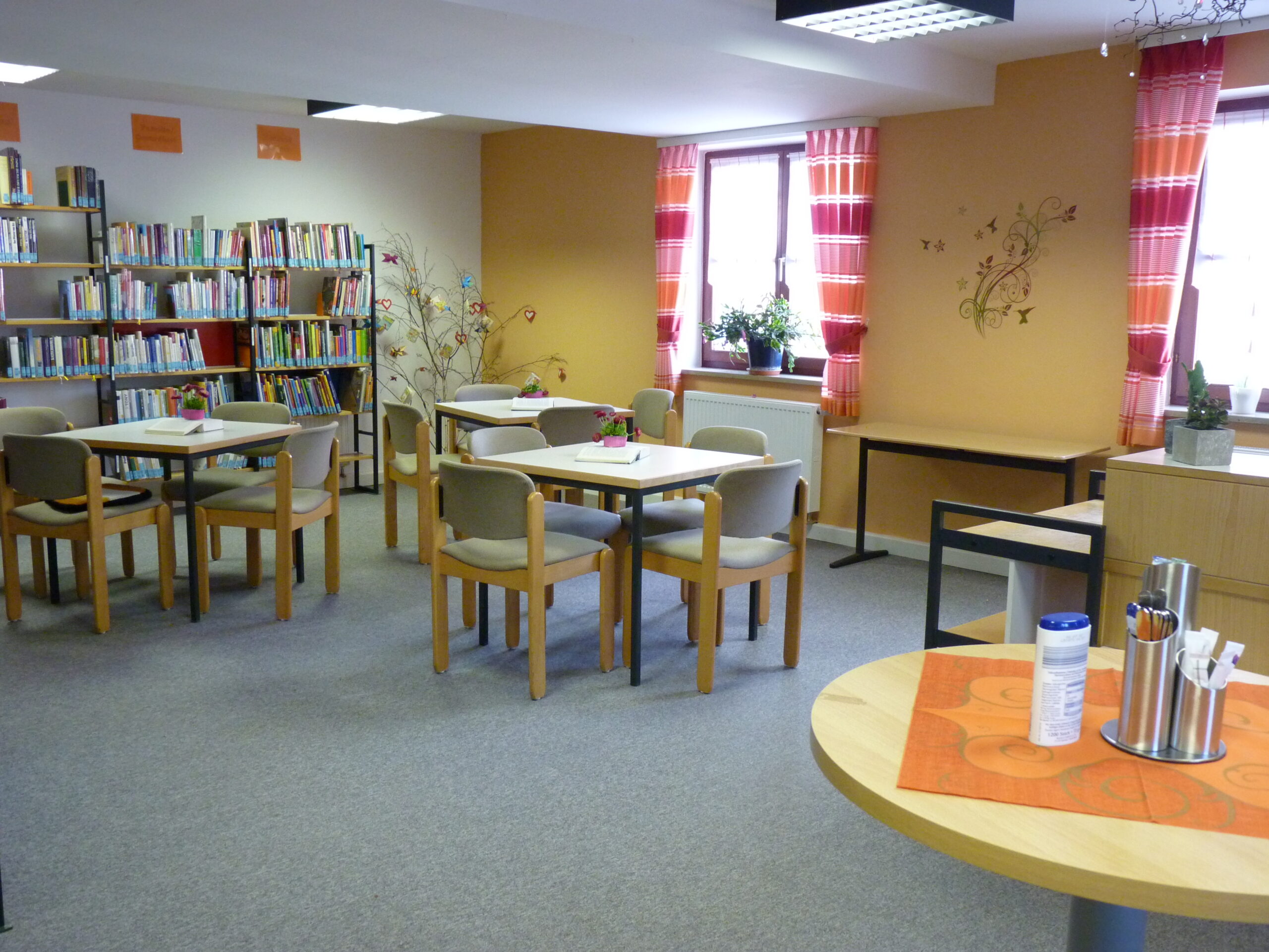 Herzlich Willkommen in der Gemeinde- und Pfarrbücherei Moosbach Raum der Bücherei mit Tischgruppen und Bücherregalen